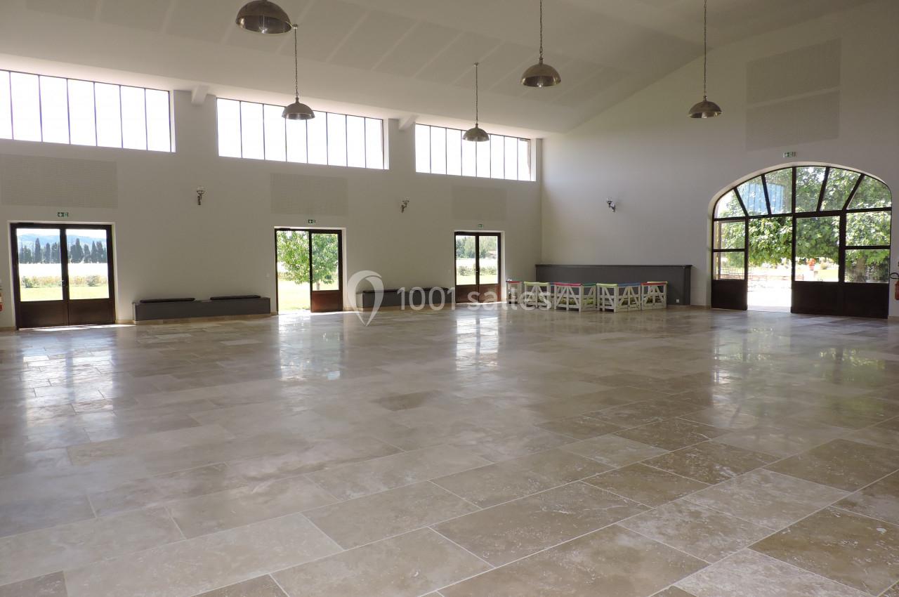 Salle spacieuse avec sol en pierre, grandes fenêtres laissant entrer la lumière et tables au fond près d'une porte voûtée.