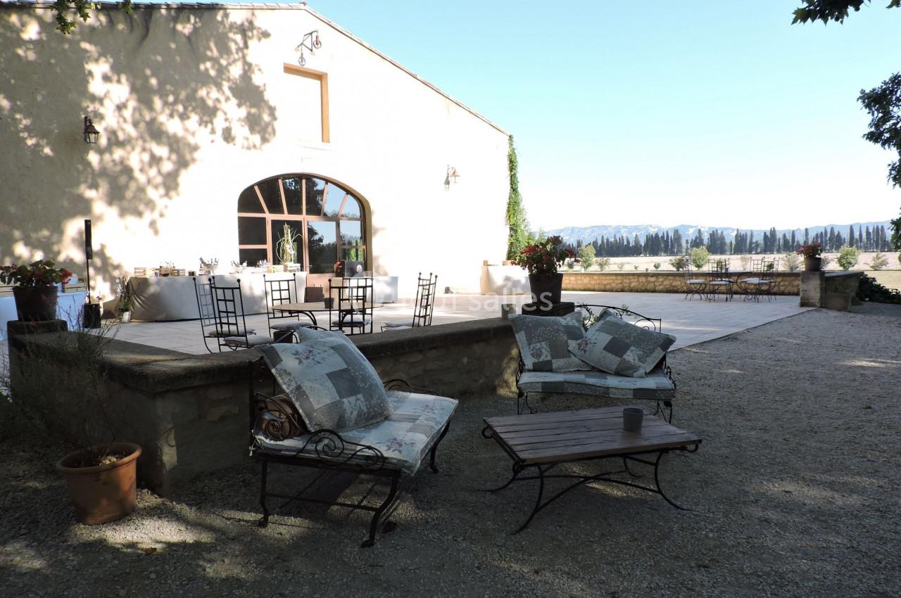 Terrasse ensoleillée avec mobilier de jardin, vue sur un paysage rural et bâtiment en pierre avec grandes fenêtres.