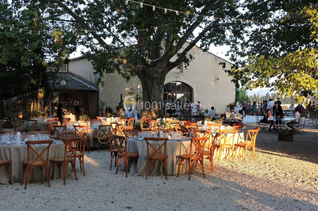 Tables dressées pour un événement en plein air sous des arbres, avec des guirlandes lumineuses et un bâtiment en arrière…
