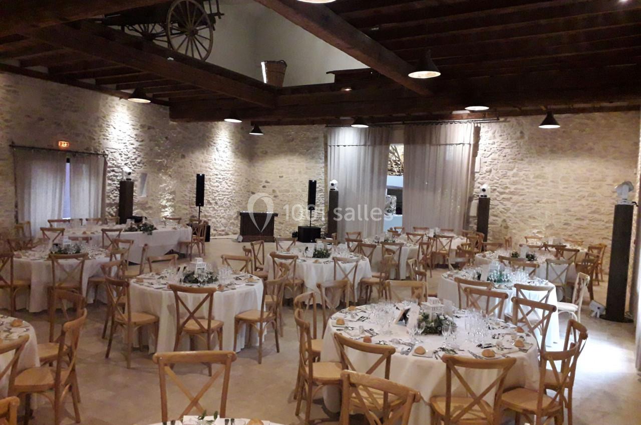 Salle de réception avec murs en pierre, poutres apparentes et tables rondes dressées pour un événement.
