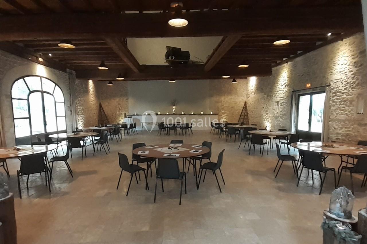 Salle spacieuse avec murs en pierre, tables rondes dressées et éclairage chaleureux, prête pour un événement.