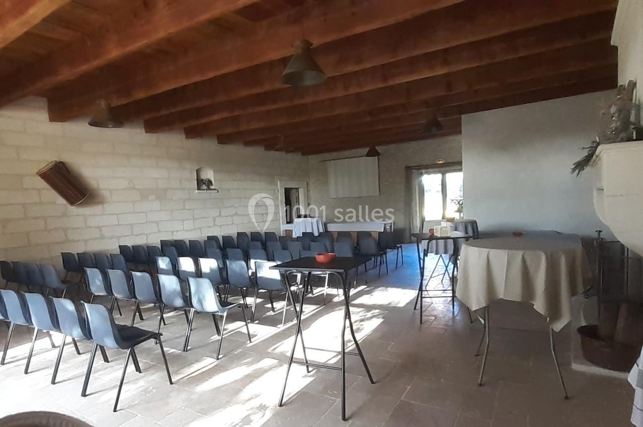 Salle lumineuse avec des rangées de chaises en plastique noir, tables recouvertes de nappes et plafond en bois apparent.