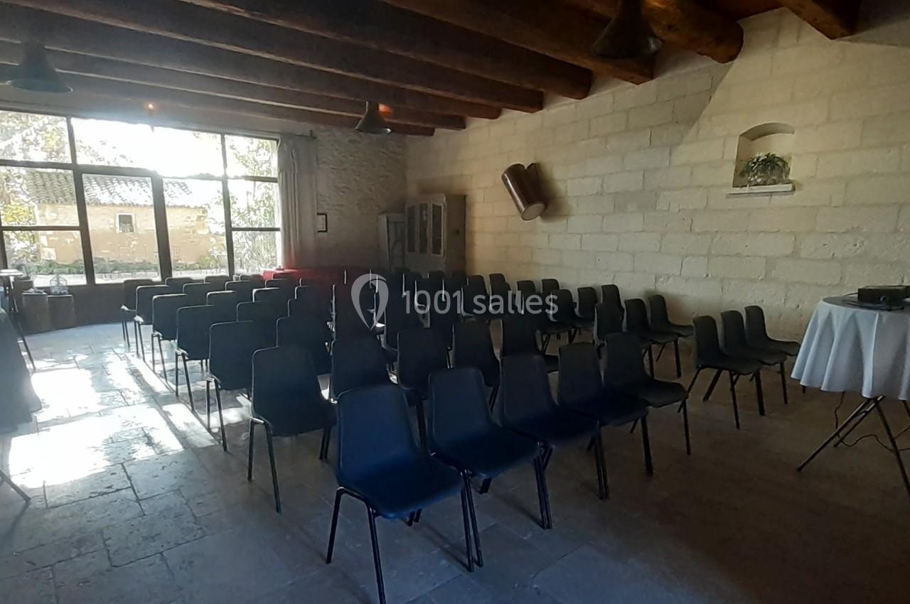 Salle avec des rangées de chaises noires vides, éclairée par une grande fenêtre donnant sur un extérieur arboré.