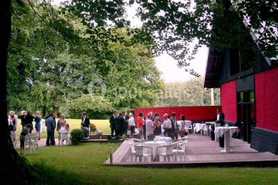 Terrasse en bois longeant un bâtiment en briques avec vue sur un jardin arboré sous un ciel bleu.