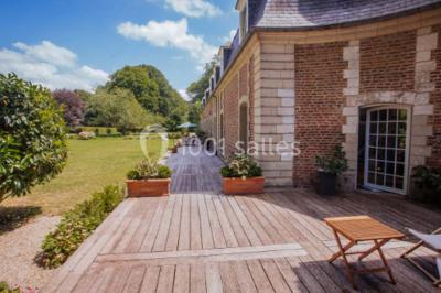 Terrasse en bois longeant un bâtiment en briques avec vue sur un jardin arboré sous un ciel bleu.