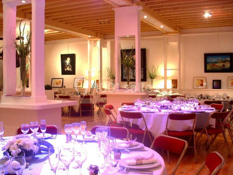 Salle lumineuse avec tables rondes dressées pour un événement, entourées de chaises rouges et décorée de tableaux.
