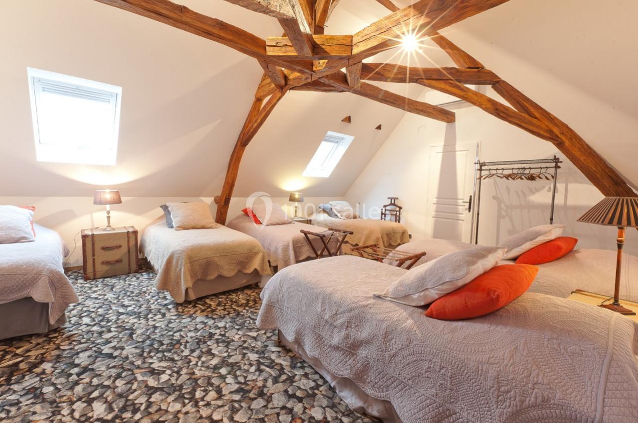 Chambre mansardée avec quatre lits simples, poutres apparentes, sol en pierre et lumière naturelle provenant des fenêtres.