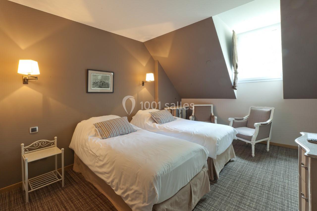 Chambre avec deux lits simples, murs beige, éclairage mural, fauteuils et fenêtre laissant entrer la lumière naturelle.