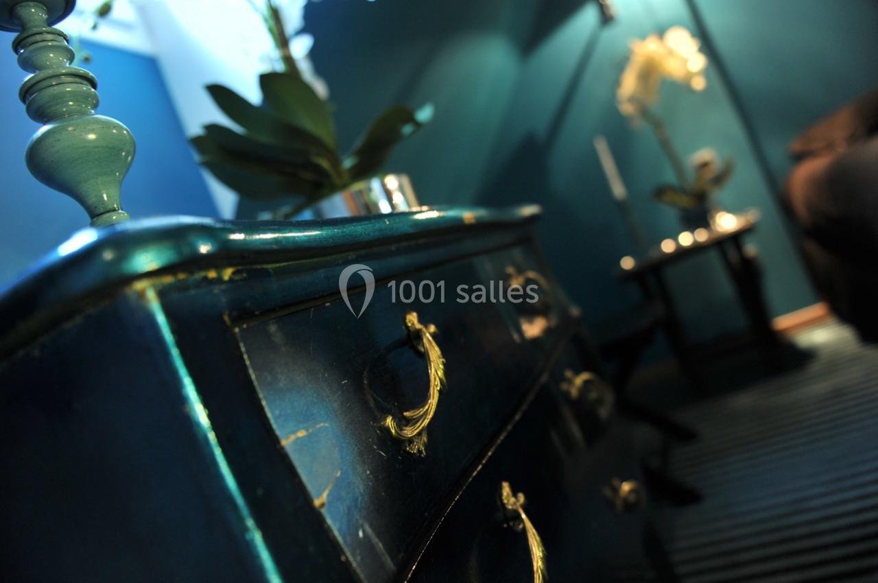 Commode en bois bleu vieilli avec poignées dorées, dans un intérieur éclairé avec des plantes et une lampe en arrière-plan.