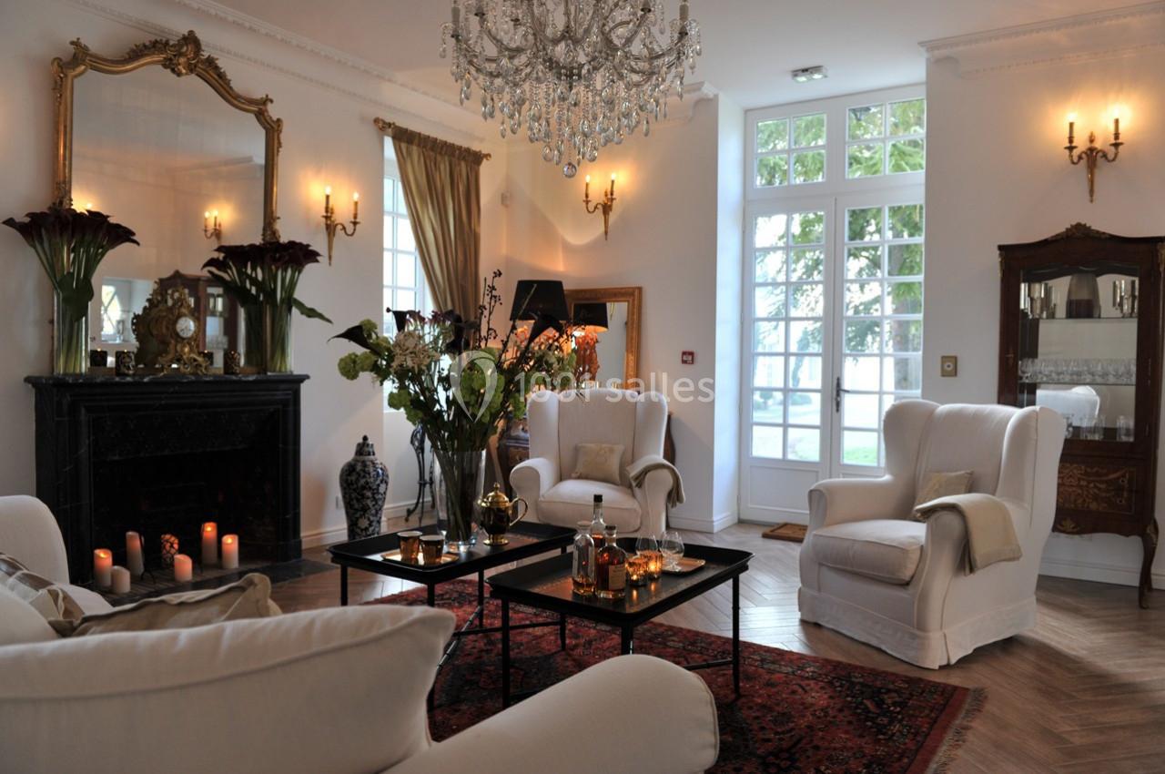 Salon élégant avec fauteuils blancs, cheminée noire ornée, lustre en cristal et grandes fenêtres laissant entrer la lumière.