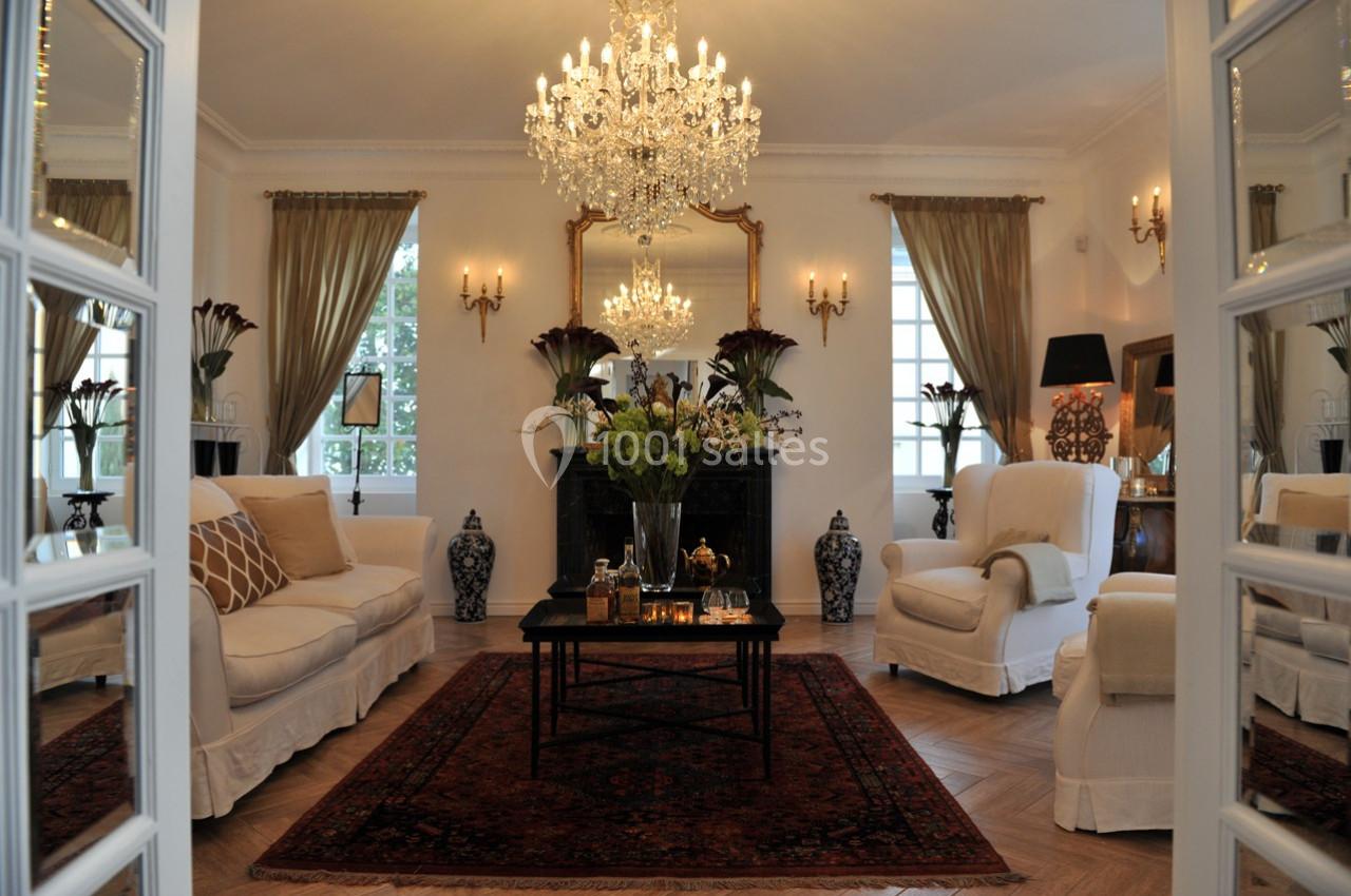Salon élégant avec lustre en cristal, meubles classiques, tapis rouge et grandes fenêtres encadrées de rideaux.
