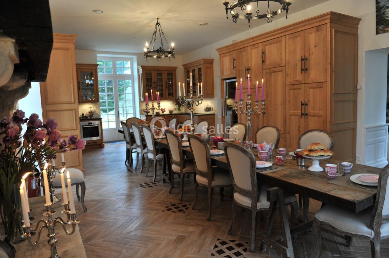 Grande cuisine lumineuse avec table en bois dressée pour un repas, entourée de chaises et décorée de chandeliers.