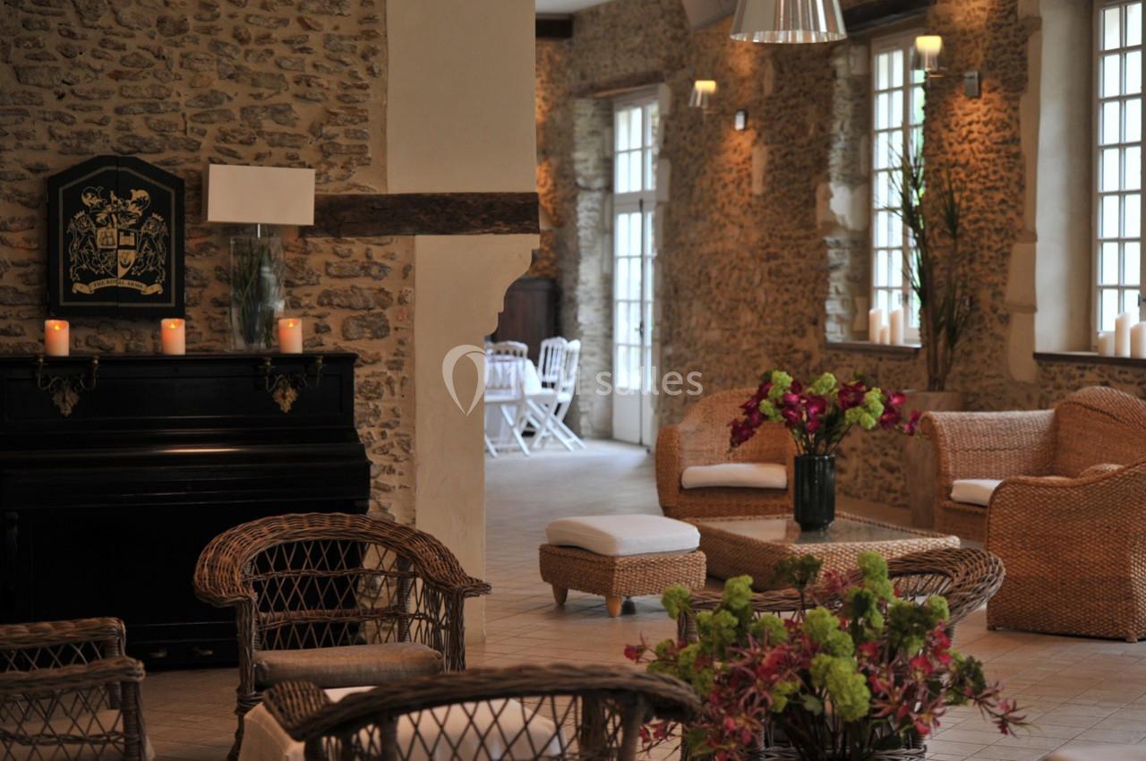Salon lumineux avec murs en pierre, mobilier en rotin, piano noir et fleurs décoratives sur une table basse.