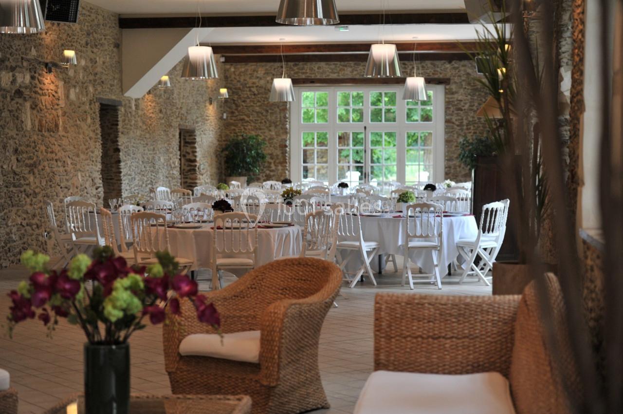 Salle de réception lumineuse avec murs en pierre, tables dressées et chaises blanches, vue sur un jardin à travers de…