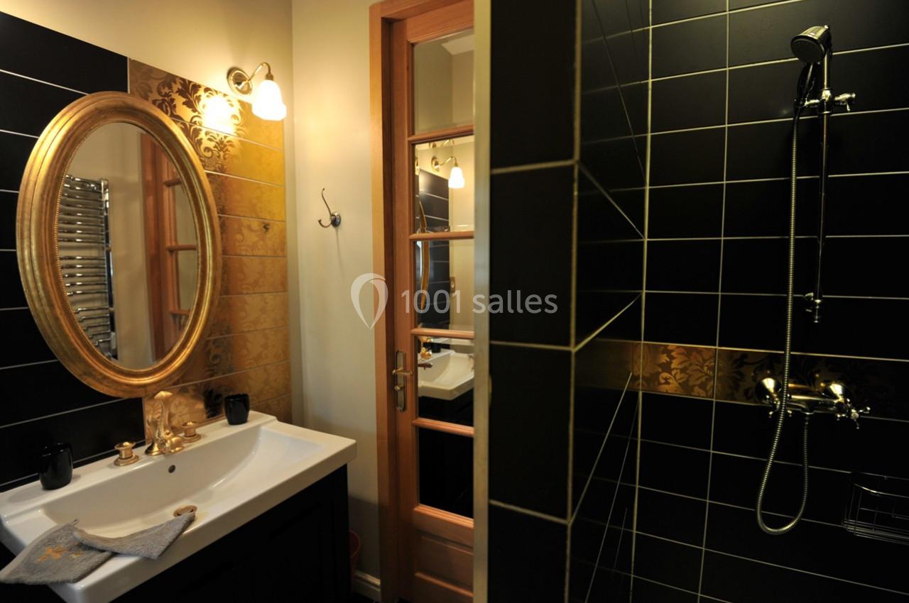 Salle de bain avec lavabo, miroir ovale doré, carrelage noir et doré, et une douche avec robinetterie murale.