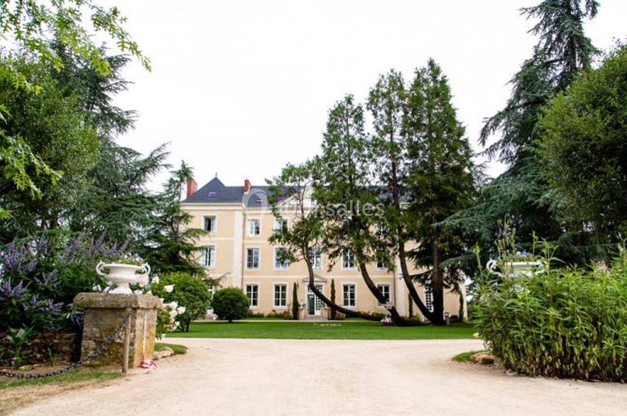 Façade d'un grand bâtiment beige entouré d'arbres et de végétation, avec une allée en gravier au premier plan.