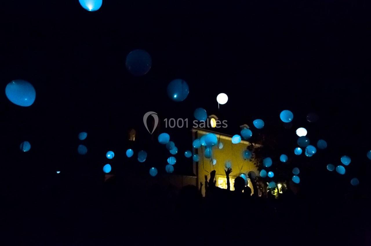 Des ballons lumineux bleus s'élèvent dans le ciel nocturne, entourés de silhouettes sombres et d'un bâtiment éclairé.