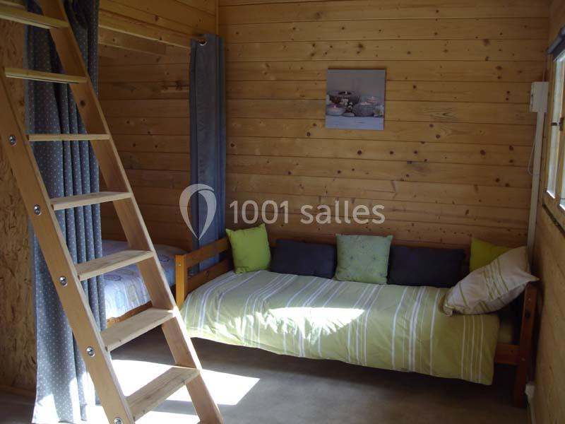 Intérieur d'une petite chambre en bois avec un lit simple, des coussins colorés et une échelle menant à une mezzanine.
