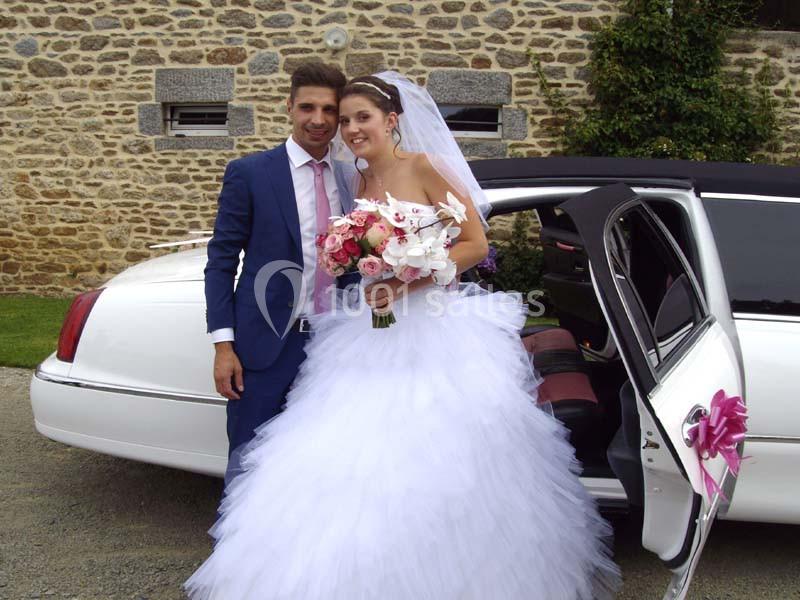 Un couple en tenue de mariage pose devant une limousine blanche garée près d'un mur en pierre.