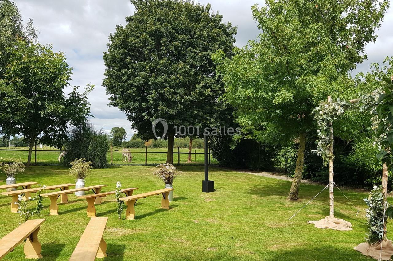 Espace extérieur aménagé pour une cérémonie avec bancs en bois, arche décorée et pelouse entourée d'arbres.