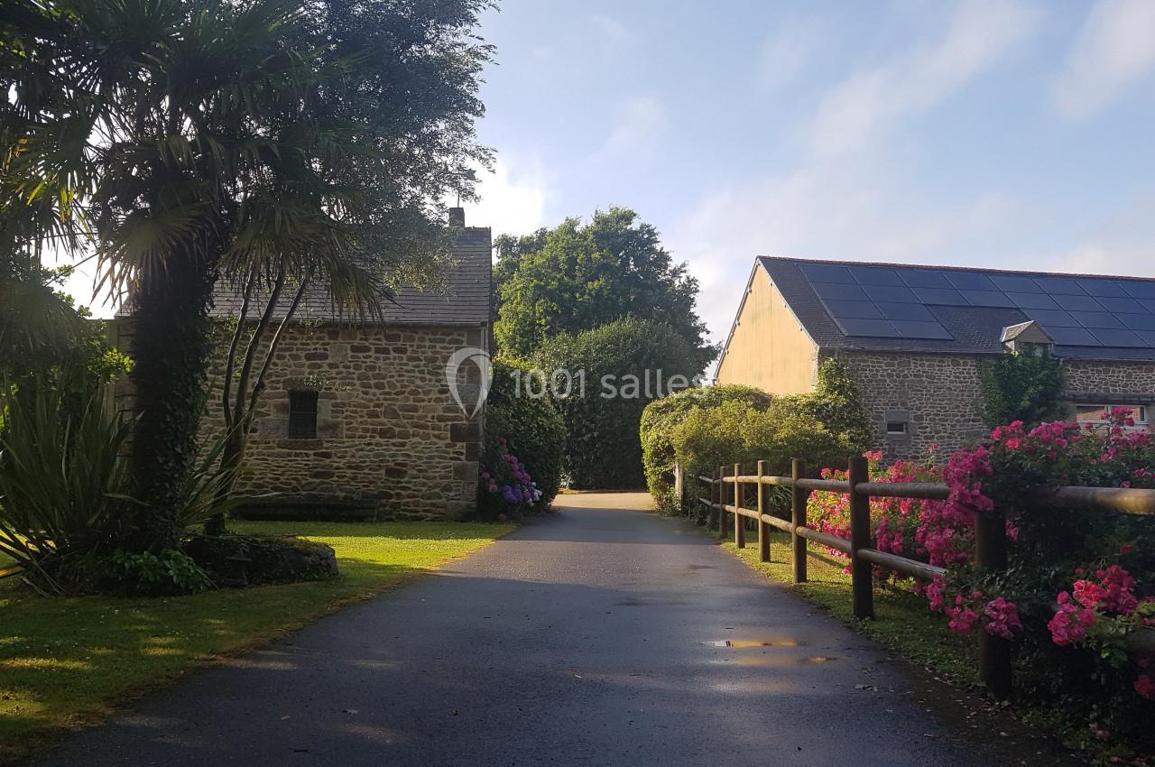 Chemin pavé bordé de fleurs et d'arbustes menant à des bâtiments en pierre sous un ciel dégagé.