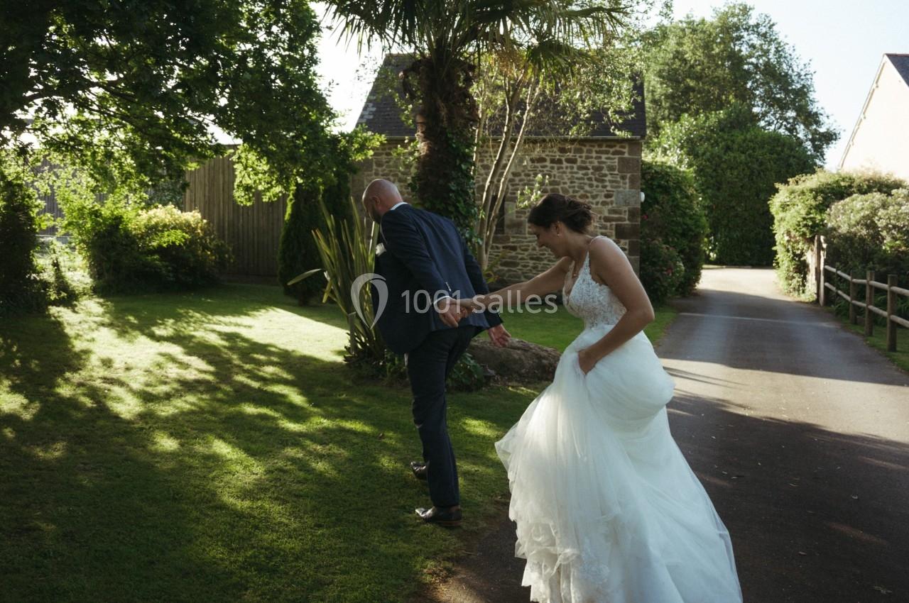 Un couple en tenue de mariage marche sur une allée bordée de verdure, éclairée par une lumière naturelle.