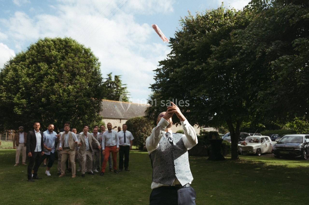 Un homme en costume lance un bouquet en l'air devant un groupe d'hommes sur une pelouse, lors d'un événement en extérieur.