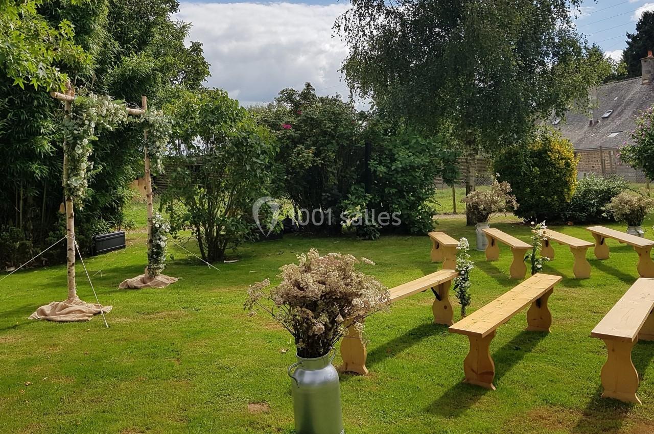 Chaises en bois disposées en extérieur devant une arche fleurie, dans un jardin verdoyant sous un ciel partiellement nuageux.