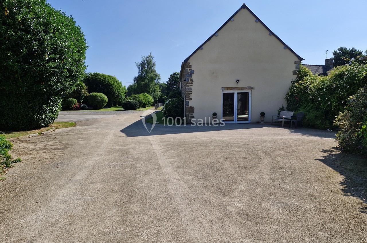 Cour en gravier devant une maison avec façade beige, entourée de végétation et d'arbres sous un ciel dégagé.