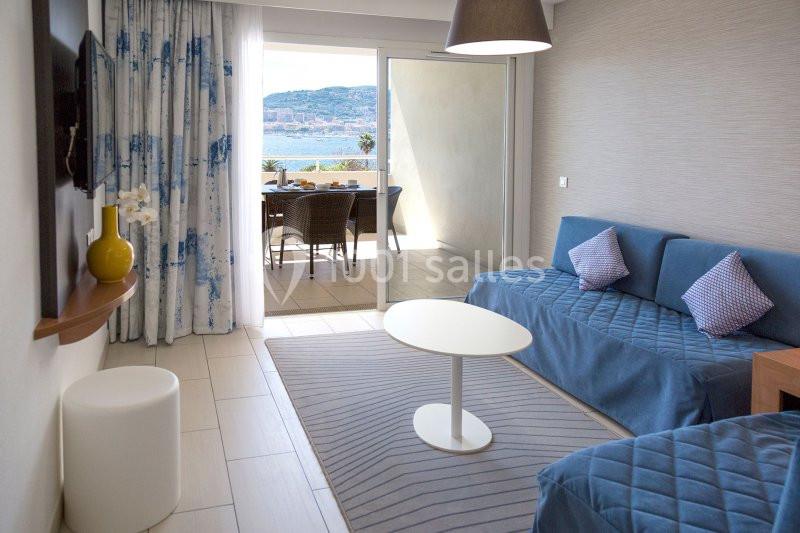 Chambre lumineuse avec deux lits, table basse, rideaux bleus, et balcon offrant une vue sur la mer et les collines.