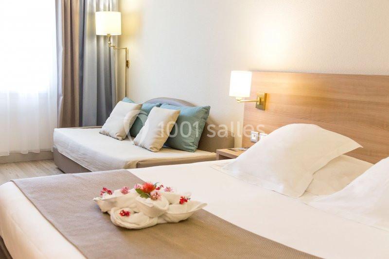 Chambre d'hôtel lumineuse avec un grand lit, un canapé-lit, des coussins colorés et une décoration florale sur le lit.