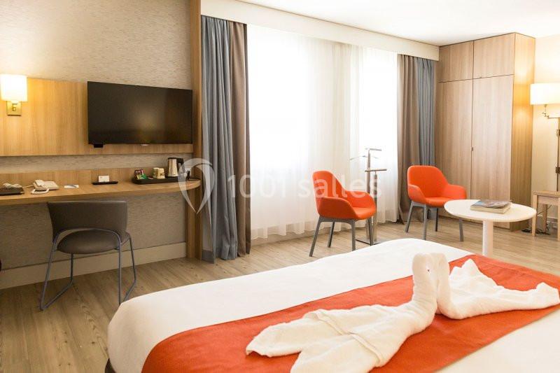 Chambre d'hôtel lumineuse avec lit, bureau, télévision, deux fauteuils orange et une table basse.