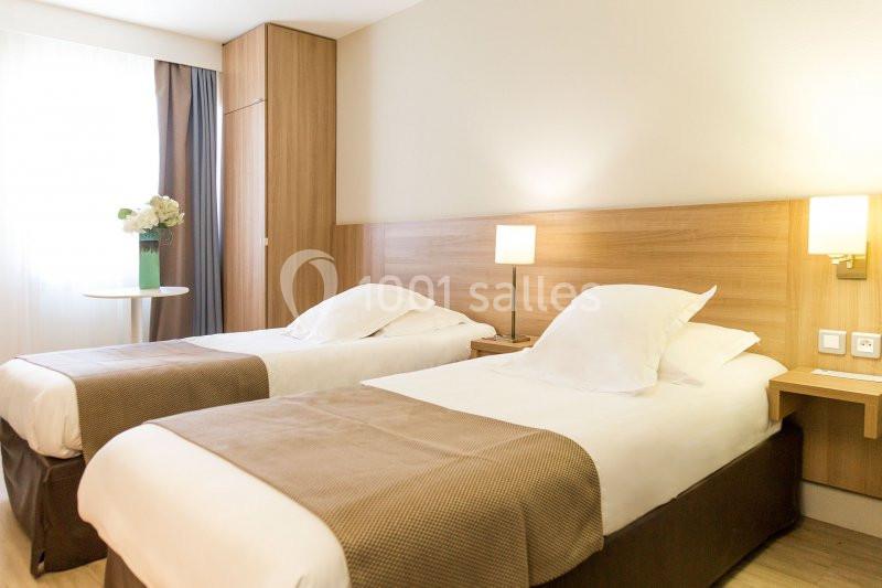 Deux lits simples avec linge blanc dans une chambre lumineuse, décorée de bois clair et équipée d'une table avec fleurs.