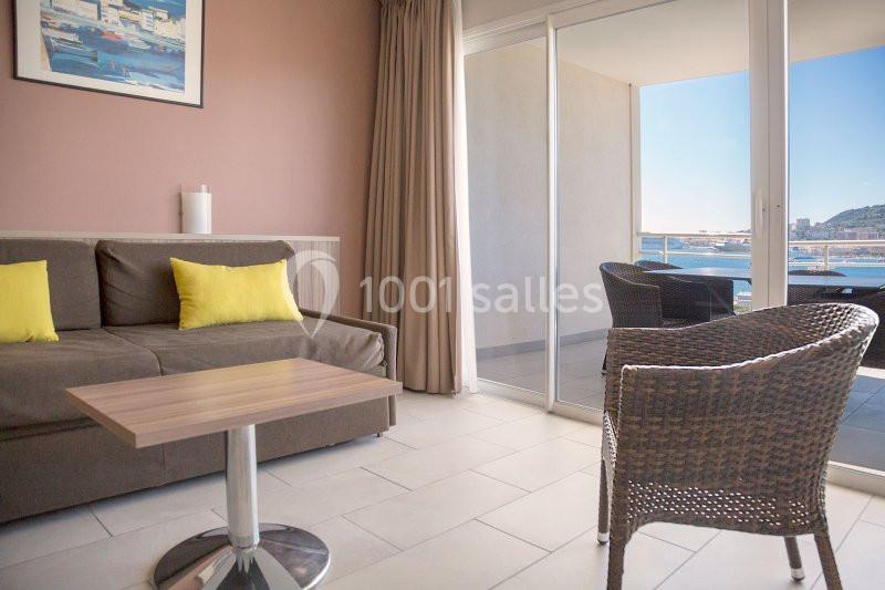 Salon lumineux avec canapé, table basse, fauteuil en rotin, baie vitrée donnant sur un balcon avec vue sur la mer.