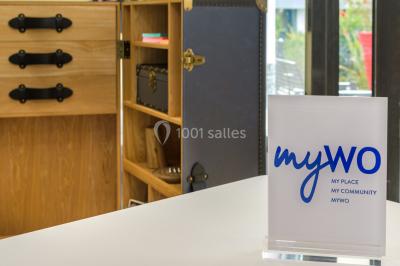 Ordinateur portable ouvert sur une table blanche avec carnet, tasse de café et plantes suspendues au-dessus.