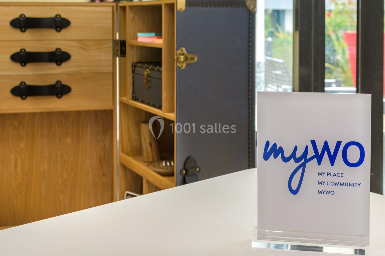Plaque avec le logo ’myWO’ posée sur une table blanche, devant un meuble en bois et une malle ouverte.