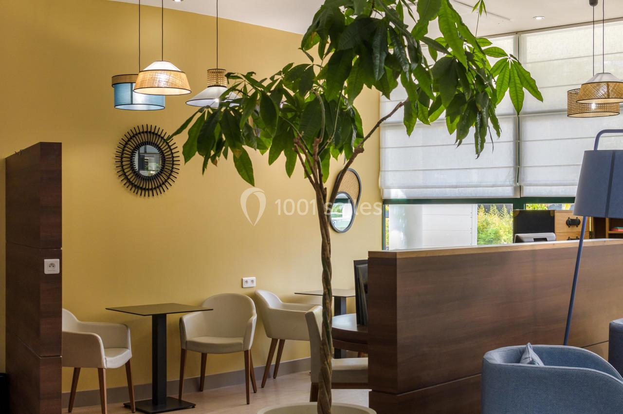 Espace intérieur lumineux avec des tables, des chaises, un grand arbre décoratif et des luminaires suspendus.