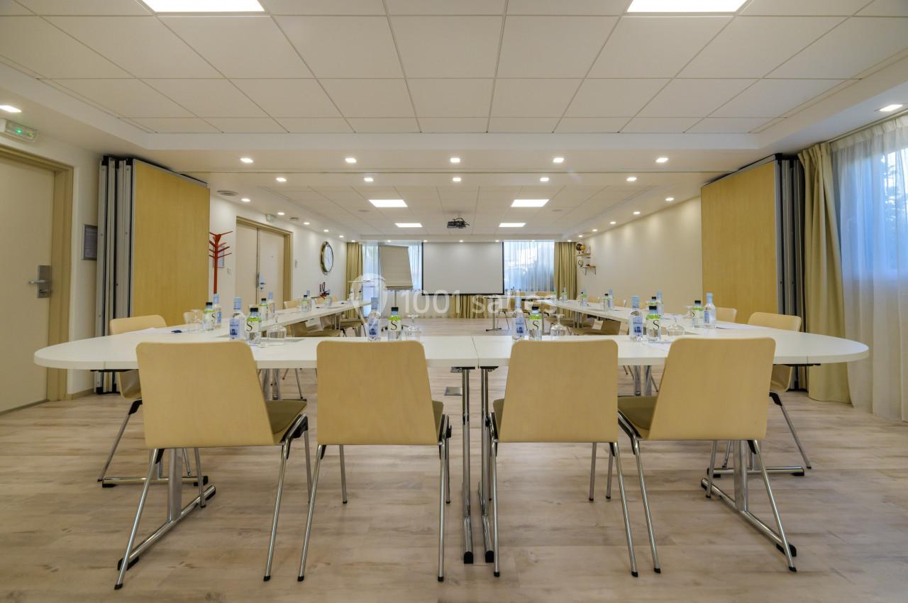 Salle de réunion lumineuse avec tables disposées en U, chaises en bois clair et écran de projection au fond.