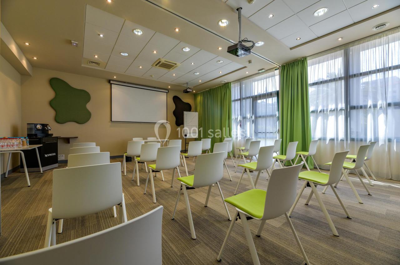 Salle de réunion lumineuse avec des chaises blanches et vertes, un écran de projection et des rideaux verts.