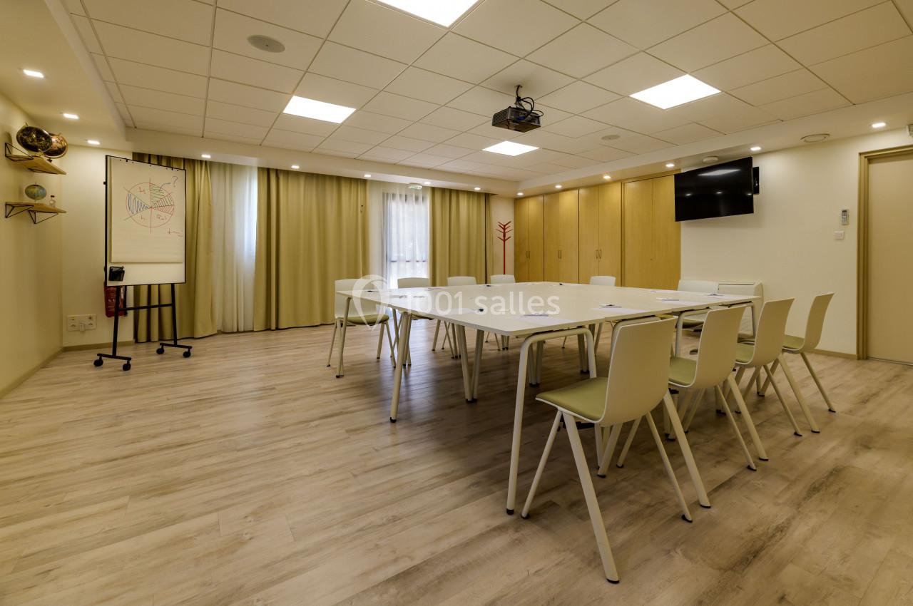 Salle de réunion lumineuse avec table rectangulaire, chaises blanches, écran mural et tableau de présentation.