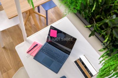 Ordinateur portable ouvert sur une table blanche avec carnet, tasse de café et plantes suspendues au-dessus.