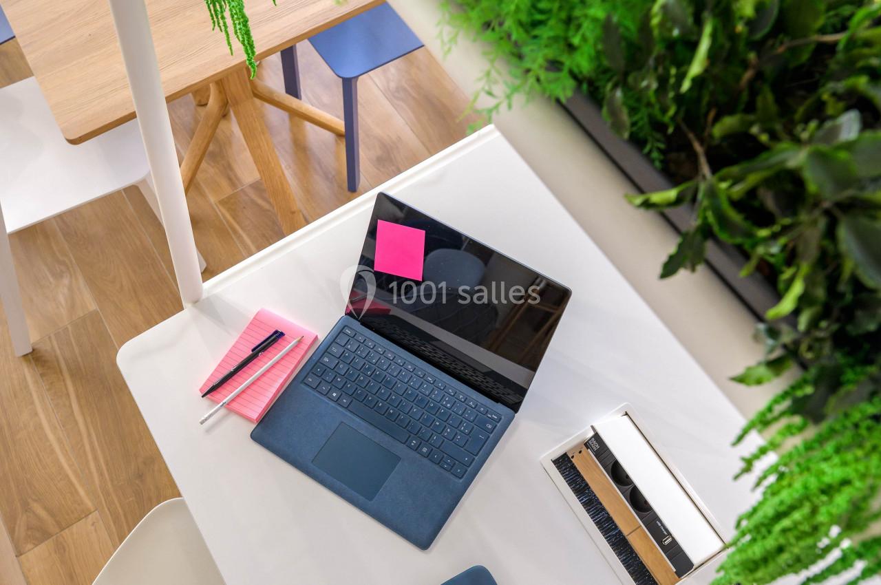 Ordinateur portable ouvert sur une table blanche avec carnet, tasse de café et plantes suspendues au-dessus.