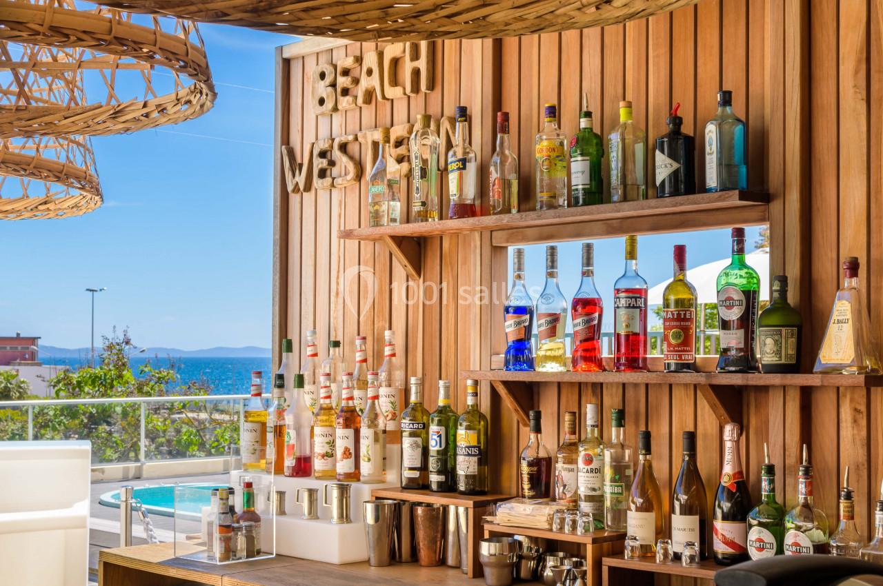 Étagères en bois garnies de bouteilles d'alcool, avec vue sur une piscine et la mer en arrière-plan.