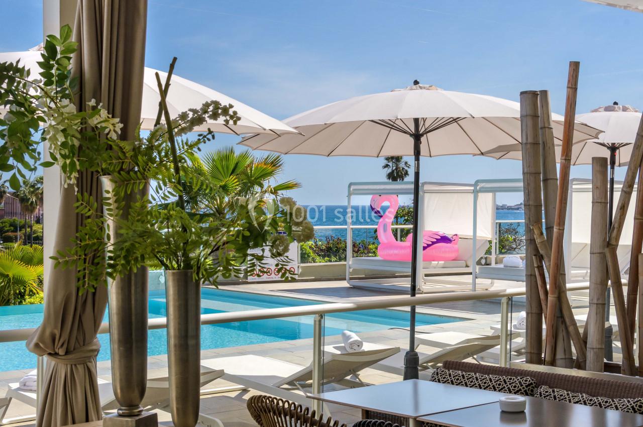 Terrasse avec vue sur une piscine, des parasols blancs, un flamant rose gonflable et la mer en arrière-plan.