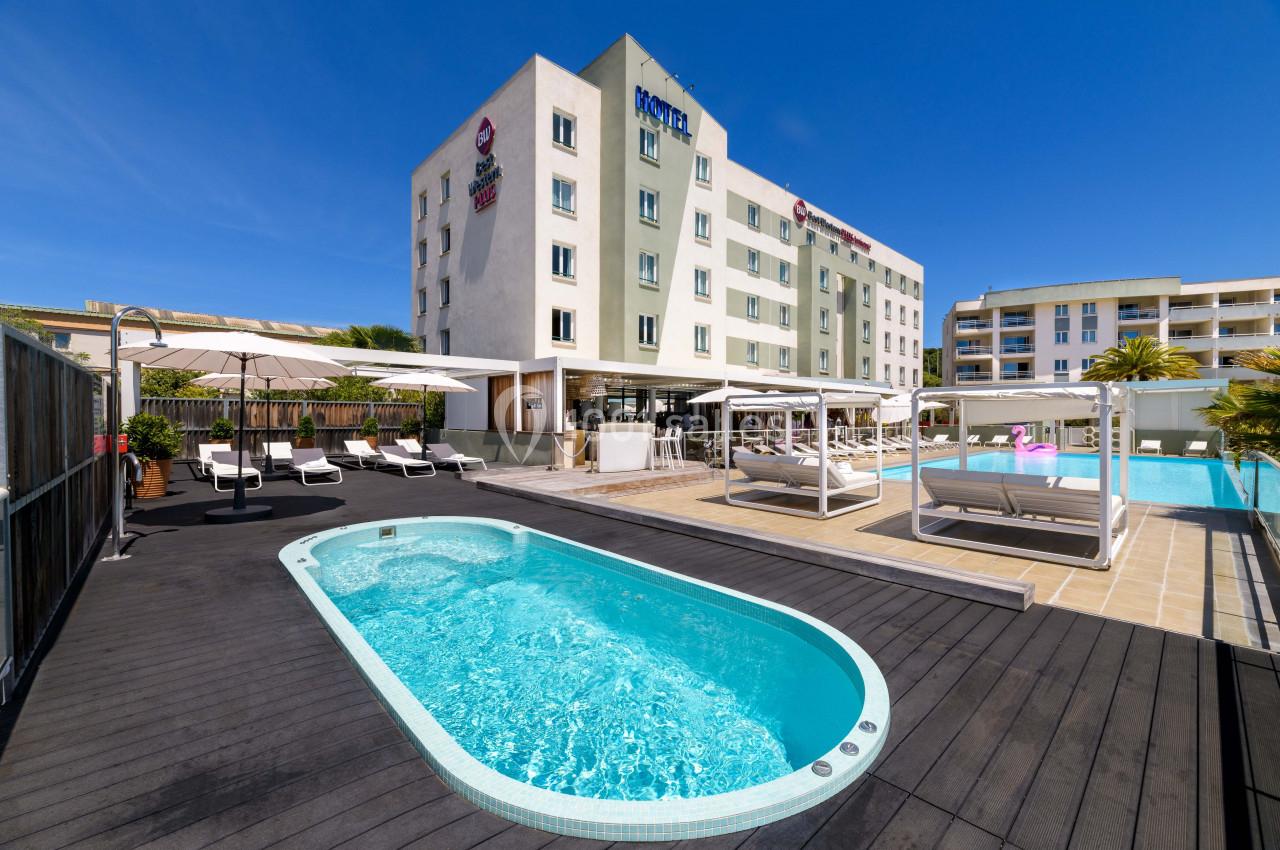 Piscine extérieure avec bain à remous entourée de transats et lits balinais, devant un hôtel moderne sous un ciel bleu.