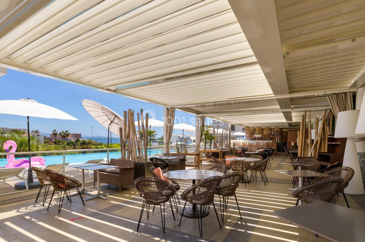Terrasse ombragée avec tables et chaises en rotin, vue sur une piscine et des parasols sous un ciel ensoleillé.