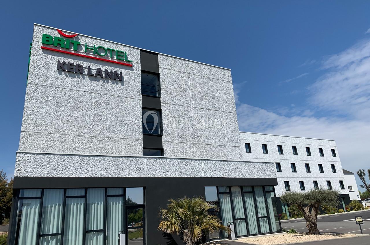 Façade d'un hôtel moderne à deux étages avec des fenêtres vitrées, des plantes et un ciel dégagé en arrière-plan.