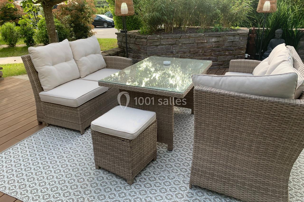 Salon de jardin en rotin avec coussins blancs, table en verre et tapis à motifs, installé sur une terrasse en bois.