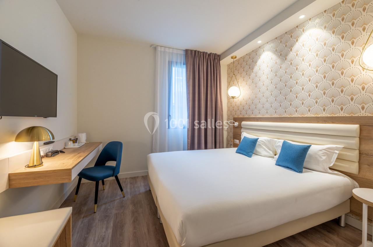 Chambre d'hôtel lumineuse avec un lit double, deux coussins bleus, un bureau, une chaise et une télévision murale.