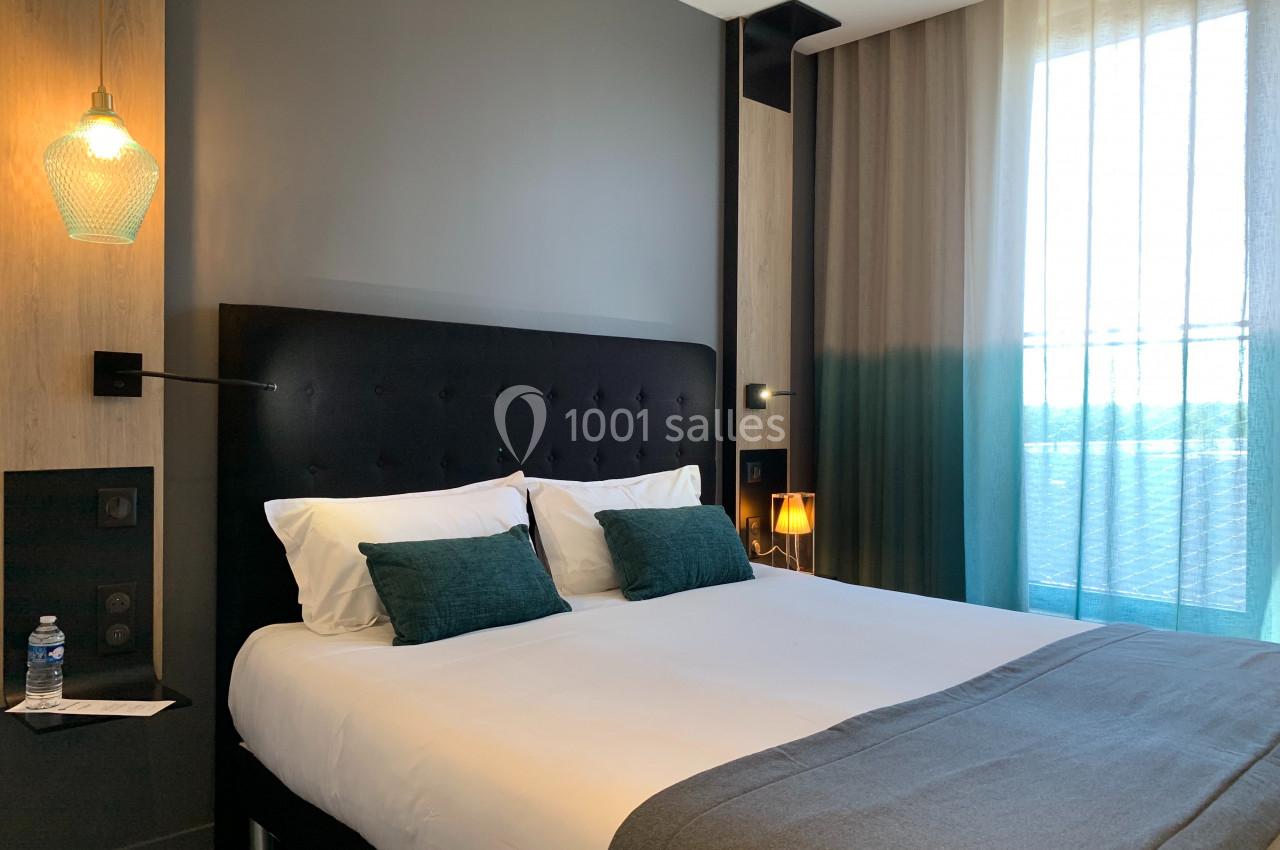 Lit double avec tête de lit capitonnée, coussins verts, éclairage doux et grande fenêtre avec rideaux.